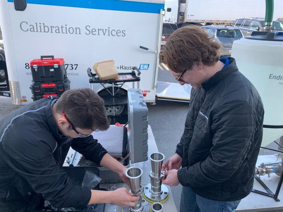 Two people in safety glasses assembling stainless sanitary fittings and an instrument head on a workbench beside a mobile calibration trailer